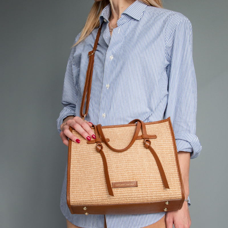 Person in a blue striped button-down shirt holding a woven straw tote bag with brown leather trim and handles.