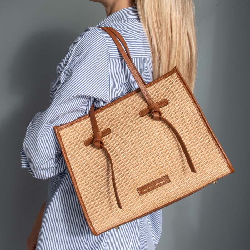 Woman wearing a light blue striped shirt carries a square tan woven tote with leather straps over her shoulder.