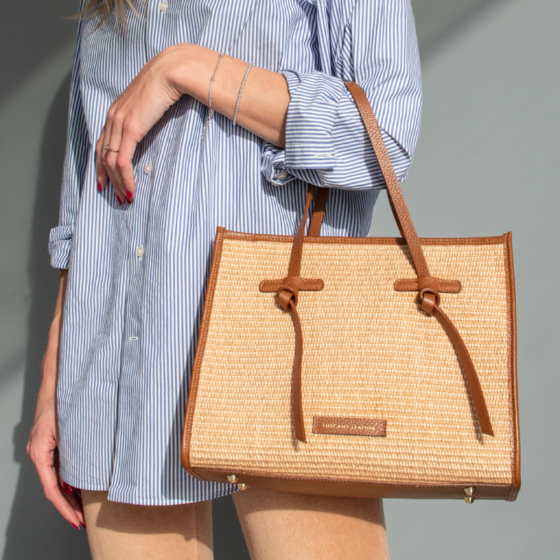 Woman in a blue striped shirt holds a woven straw tote with tan leather trim and straps.