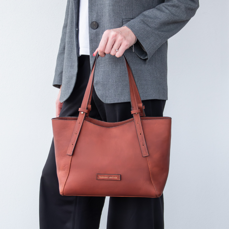 Person in a gray blazer carries a rust-brown leather tote bag by its twin handles outside a light wall backdrop, showing a small brand plaque on the bag.