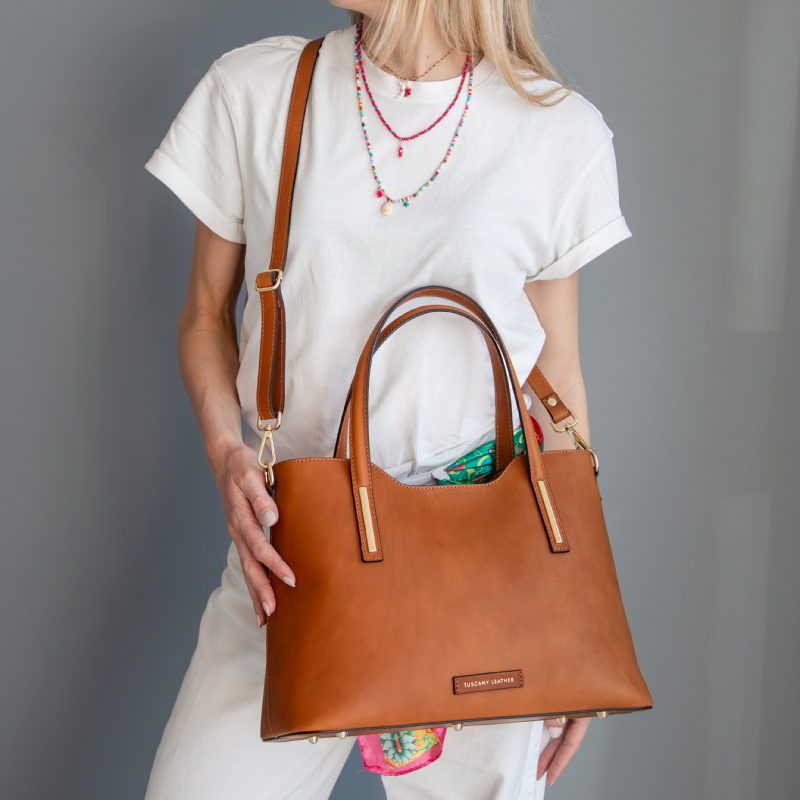 Tan leather tote with double handles and a long strap, carried by a person in a white shirt; a colorful scarf peeks from the bag opening.
