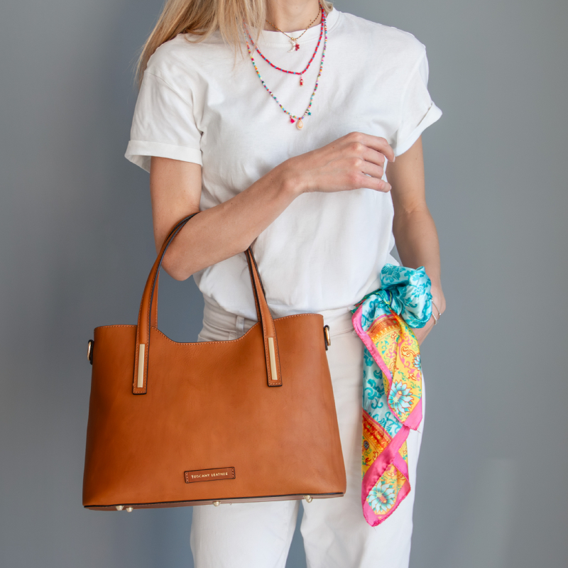 Woman in a white shirt and pants holding a tan leather tote bag with a colorful scarf tied to it.