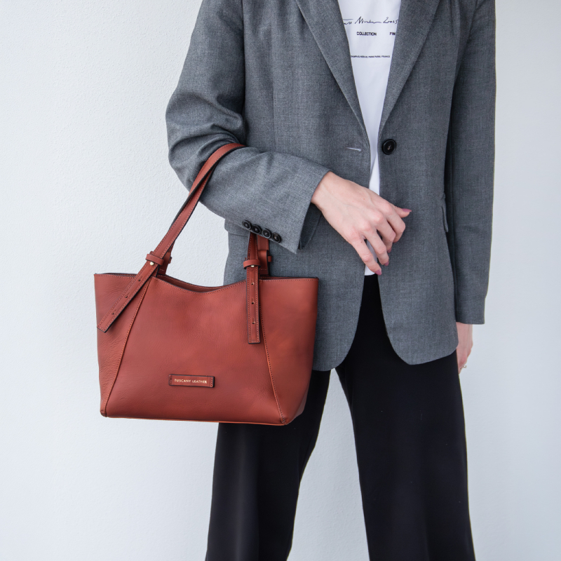 Woman in a gray blazer and black pants holding a brown leather handbag by its strap on her left side.